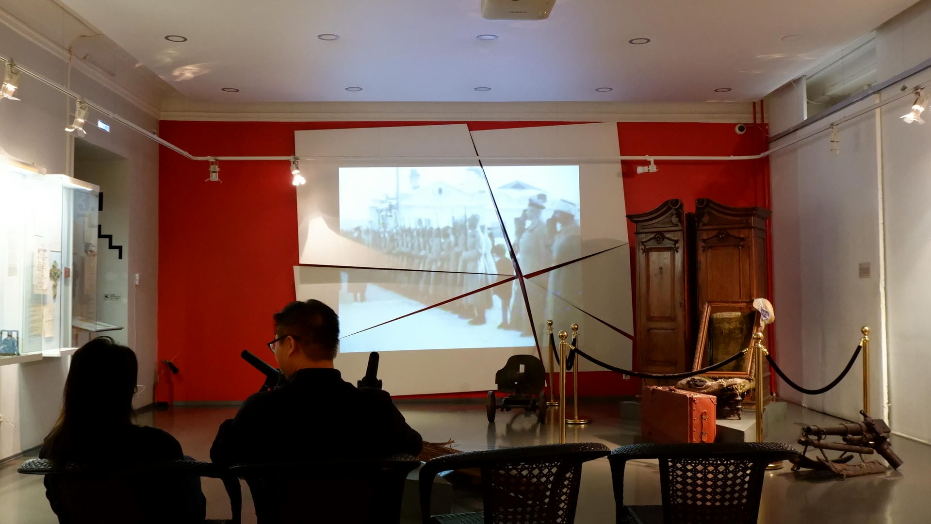 Two people sit on chairs watching historical footage projected onto the wall.