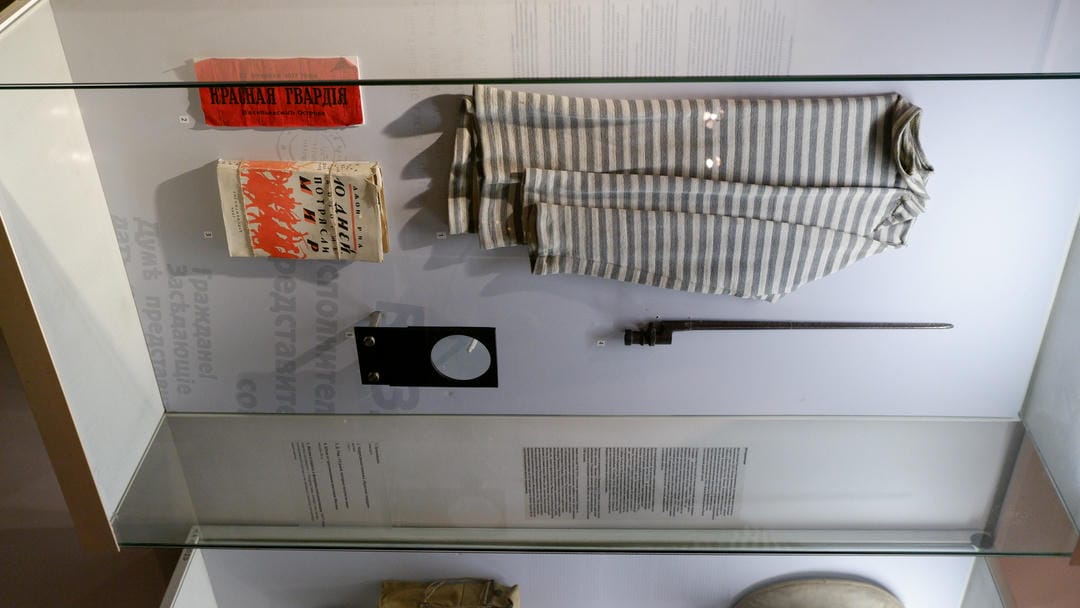 Striped prison uniform, books, and a bayonet displayed in a glass cabinet.