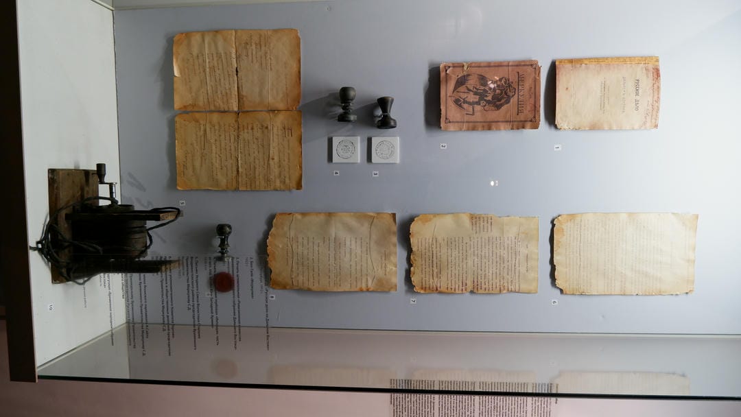 Multiple old documents and stamps displayed in a glass cabinet, with a black machine at the bottom.