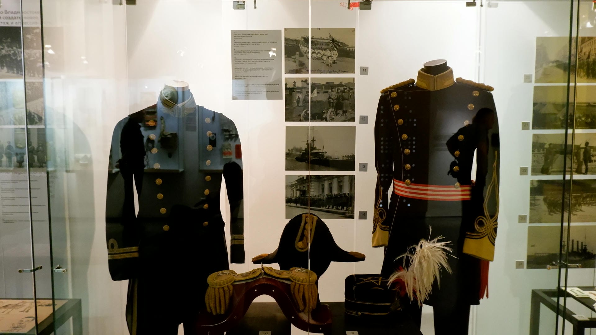 Two military uniforms and a military cap displayed in a glass cabinet, accompanied by historical photographs.