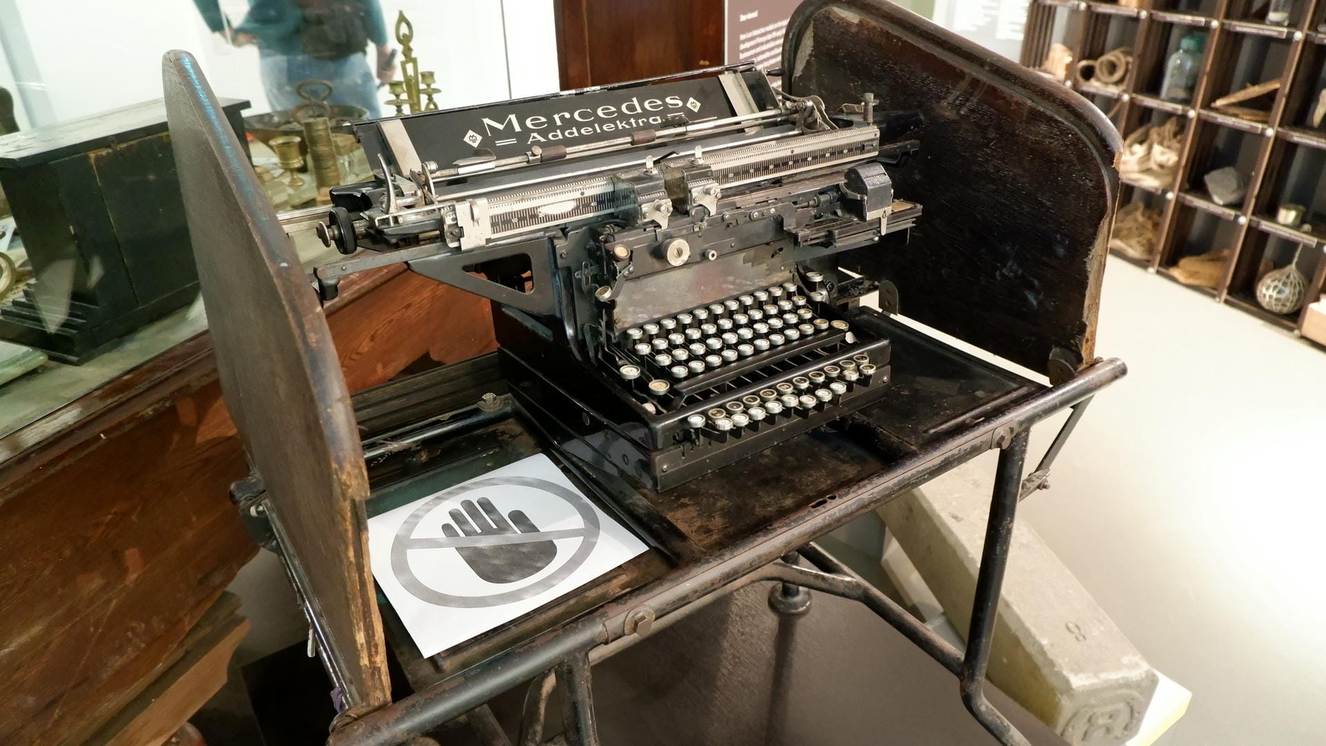 An old typewriter branded Mercedes Addelektra, placed on a wooden base, with a “Do Not Touch” sign beside it.