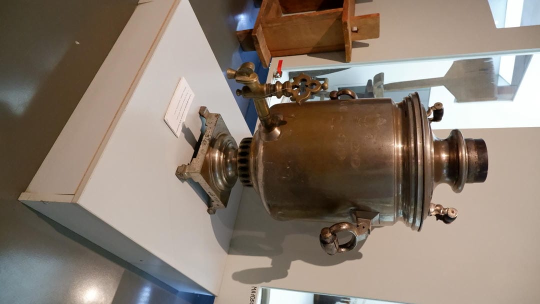 A large metal samovar with decorative handles and spout, placed on a white platform.