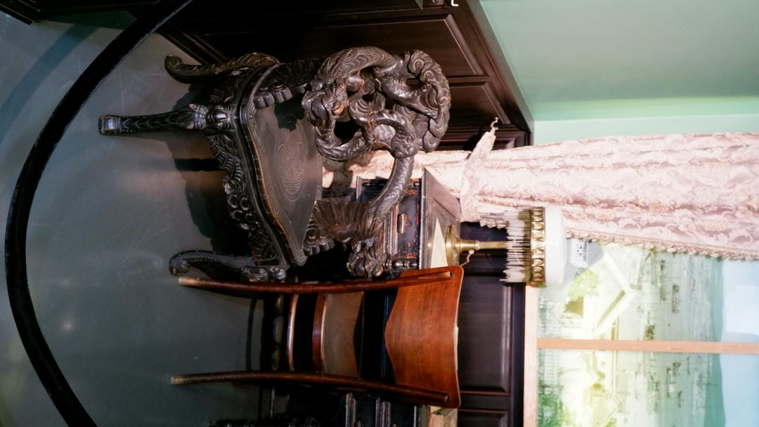 An antique chair with a carved dragon pattern on the backrest, next to a wooden table and a regular chair.