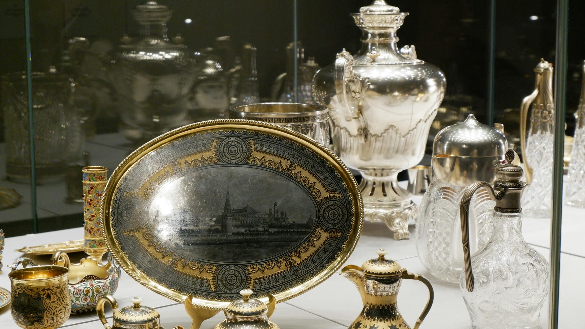 Silver cutlery and tableware, including trays, teapots, and bottles, with cityscape engravings on the trays, displayed in a showcase.