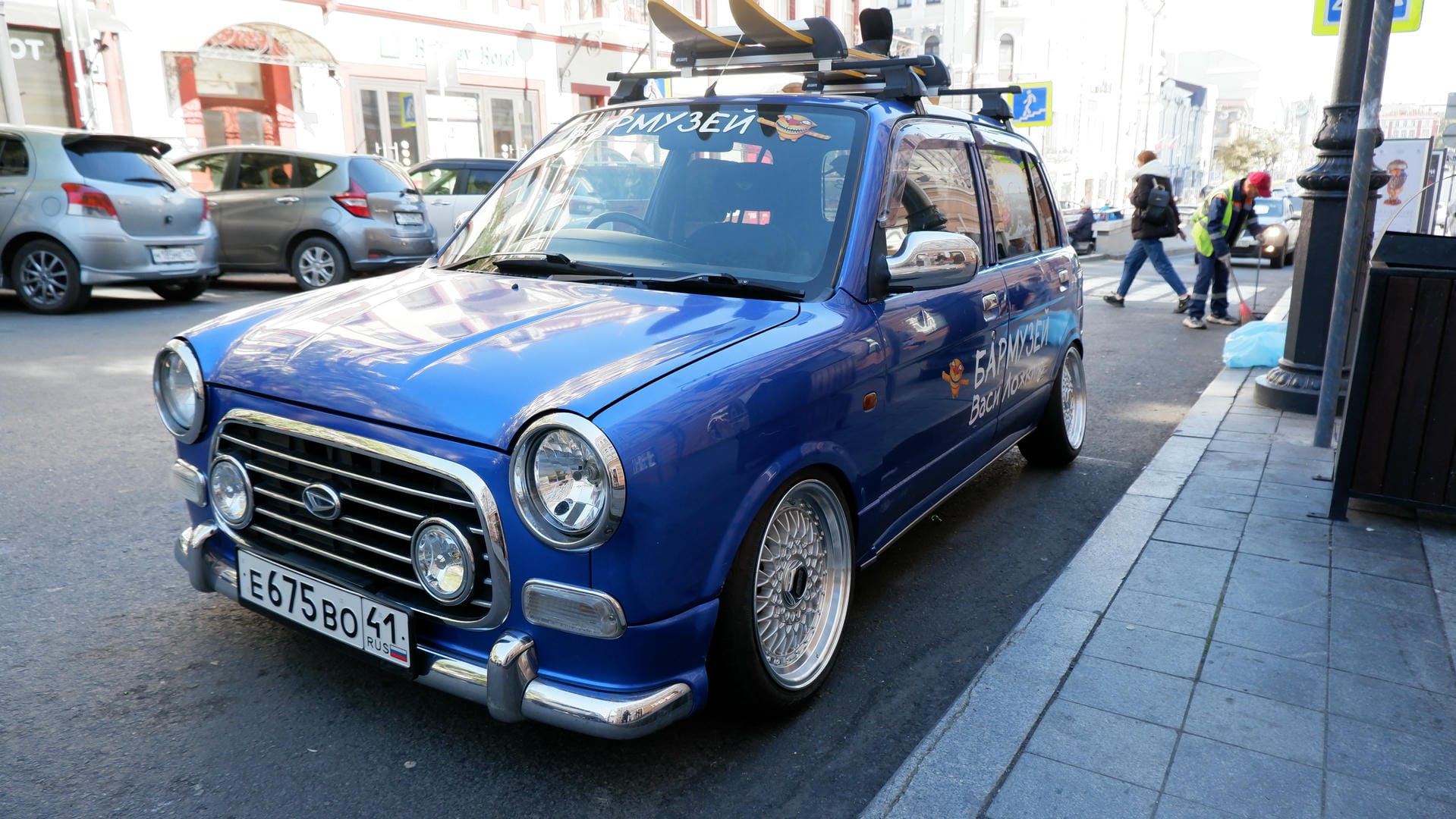 A blue car parked by the roadside with a surfboard on the roof and cartoon patterns on the body.