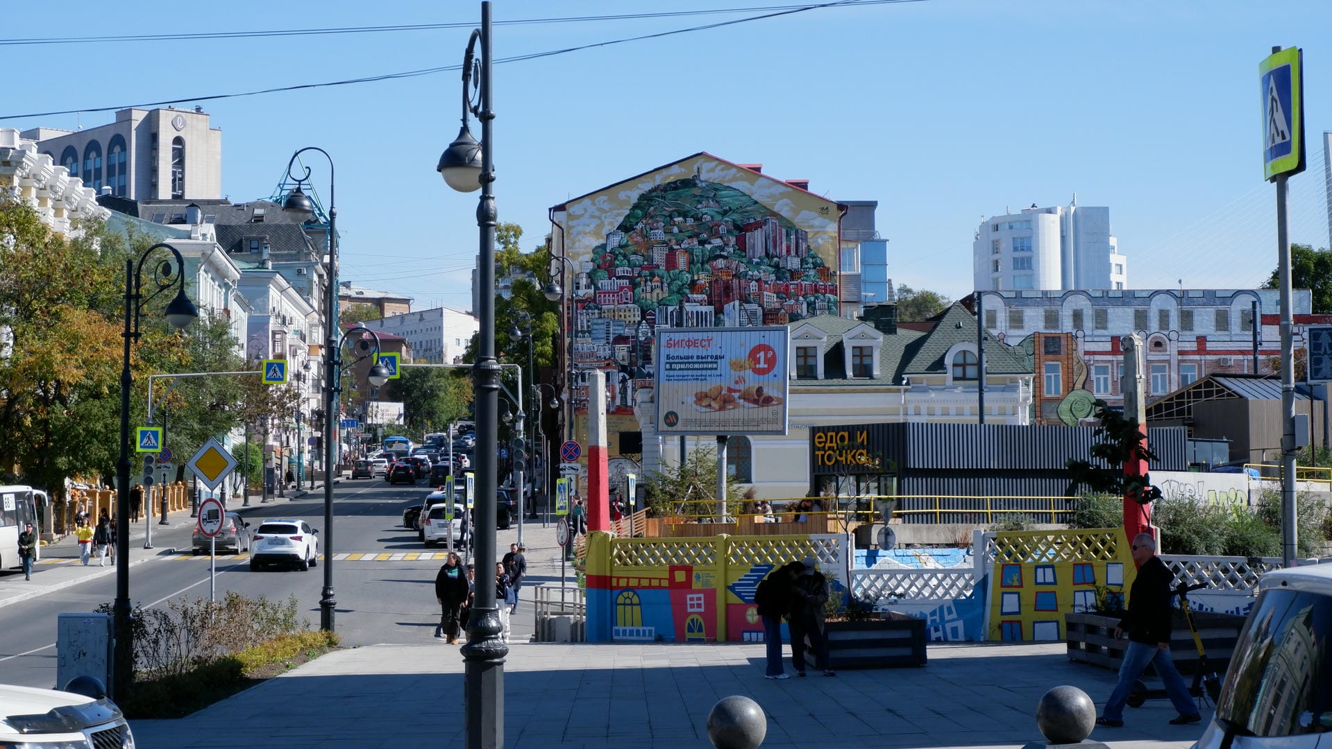 Buildings lining both sides of the street, with a large mural depicting urban scenery in the distance.