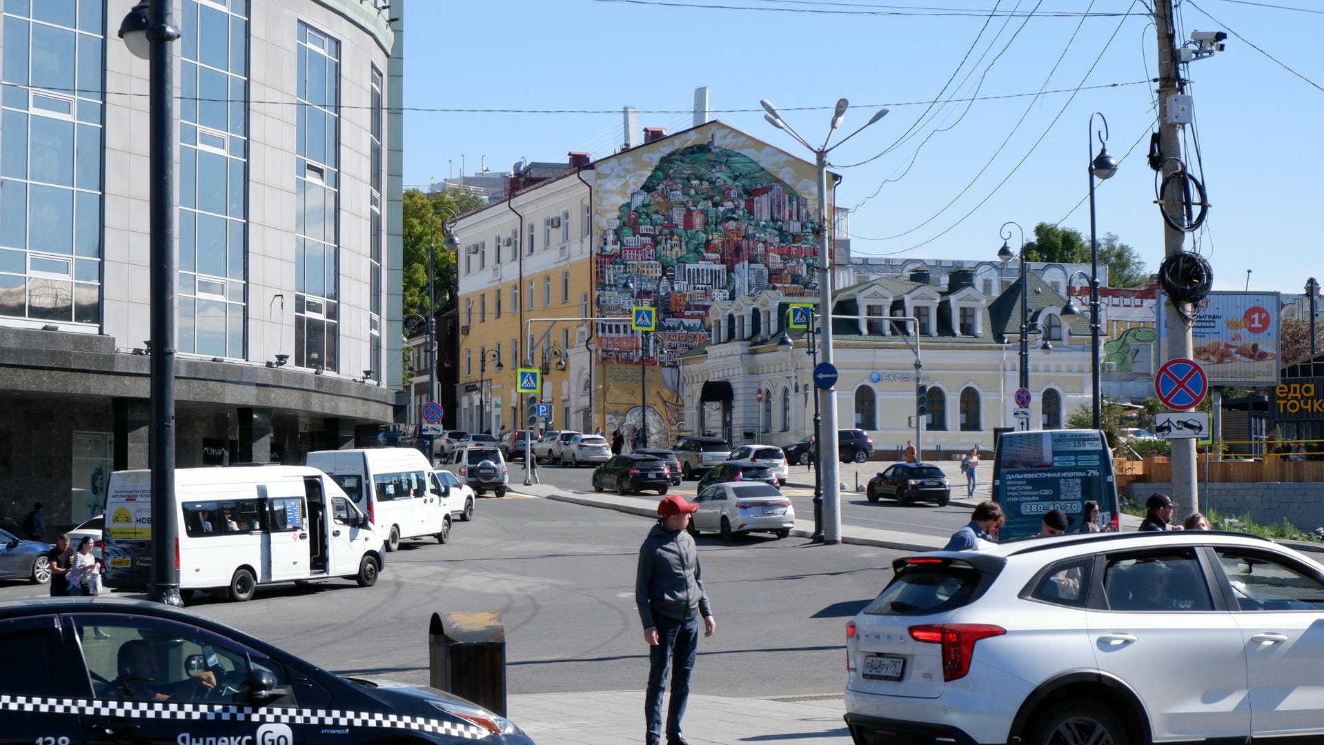 A busy city street with modern buildings on the left, old buildings decorated with large murals on the right, and pedestrians and vehicles on the road.