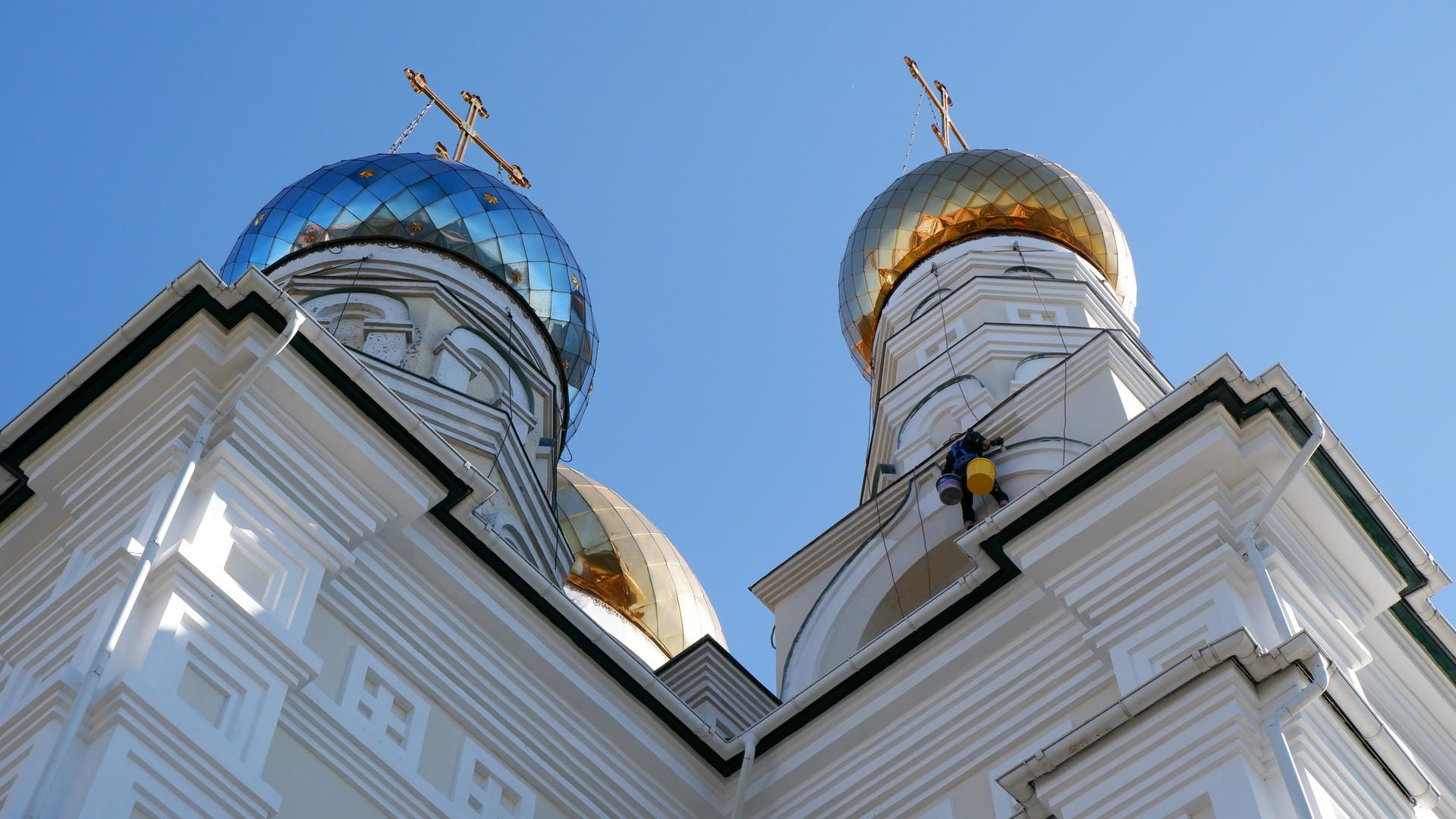 The top of a white church with two blue domes and one golden dome, all adorned with crosses, and a worker cleaning one of the domes.