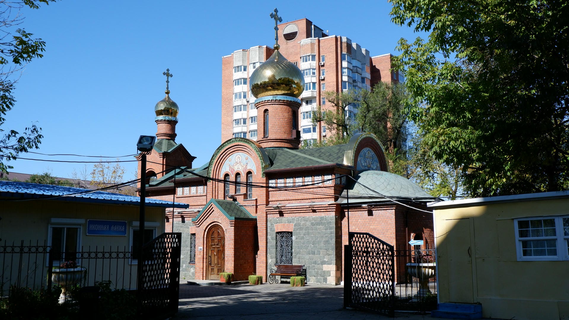 A red-brick church with a golden dome, a wrought-iron gate in front, and a high-rise building in the background.