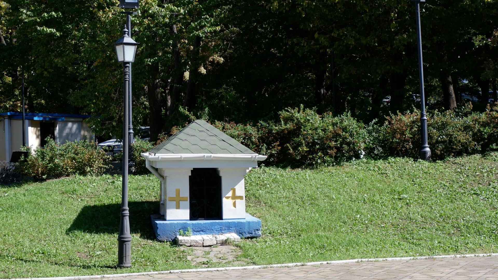 A small building in a park with yellow crosses on white walls, a green roof, and surrounded by trees and shrubs.