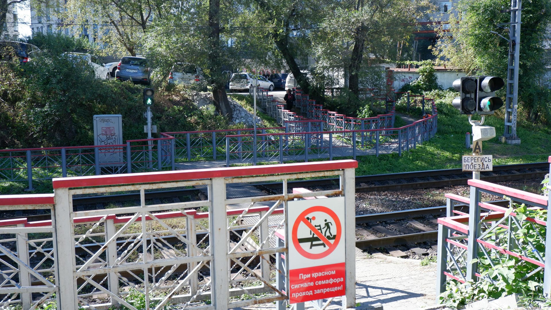 Railway tracks with red railings and warning signs, with trees and buildings in the distance.