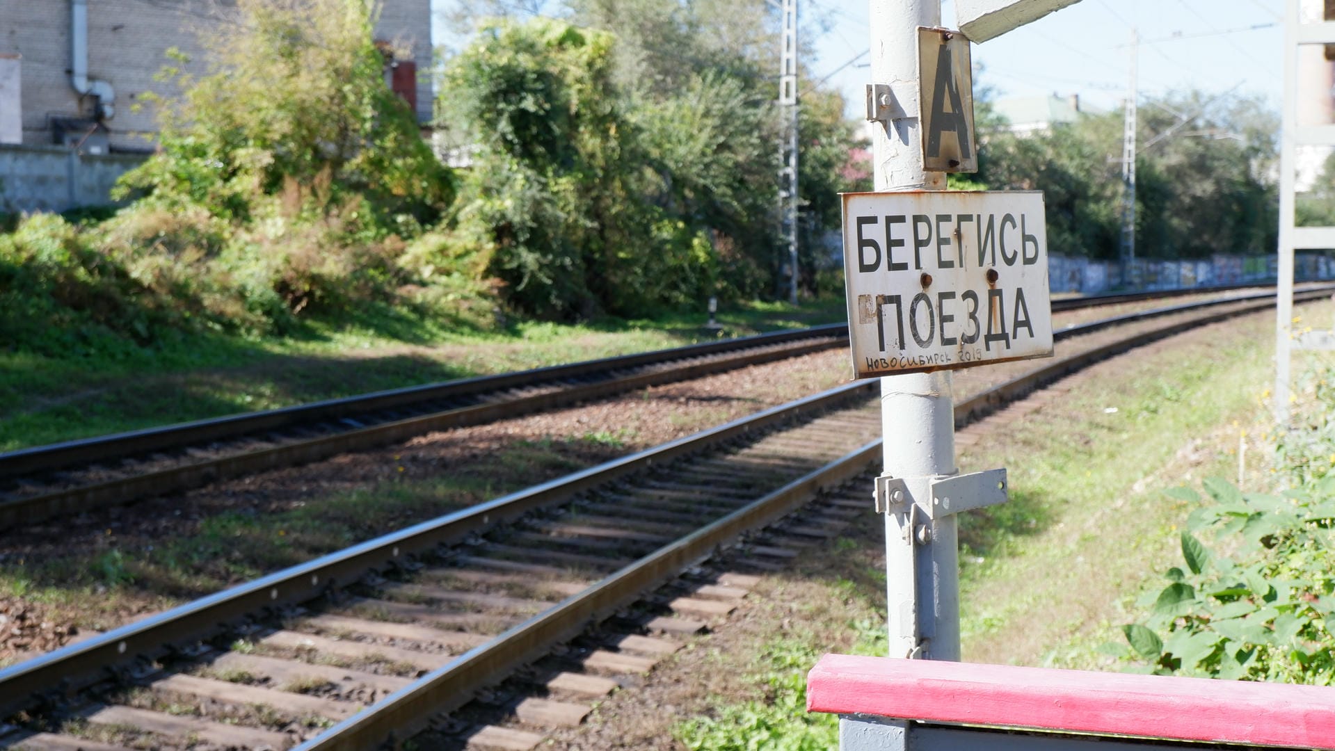 A sign reading “Caution Trains” stands beside railway tracks, with trees and utility poles in the background.