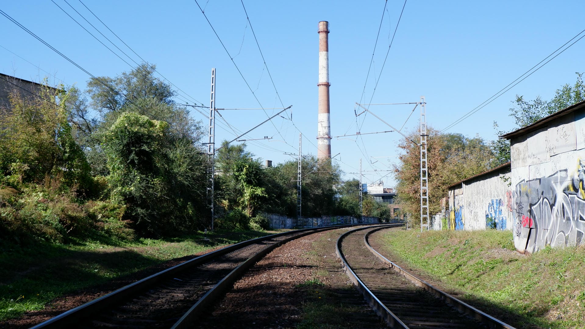 Two railway tracks extending into the distance, with trees on the left and graffiti-covered walls and a chimney on the right.