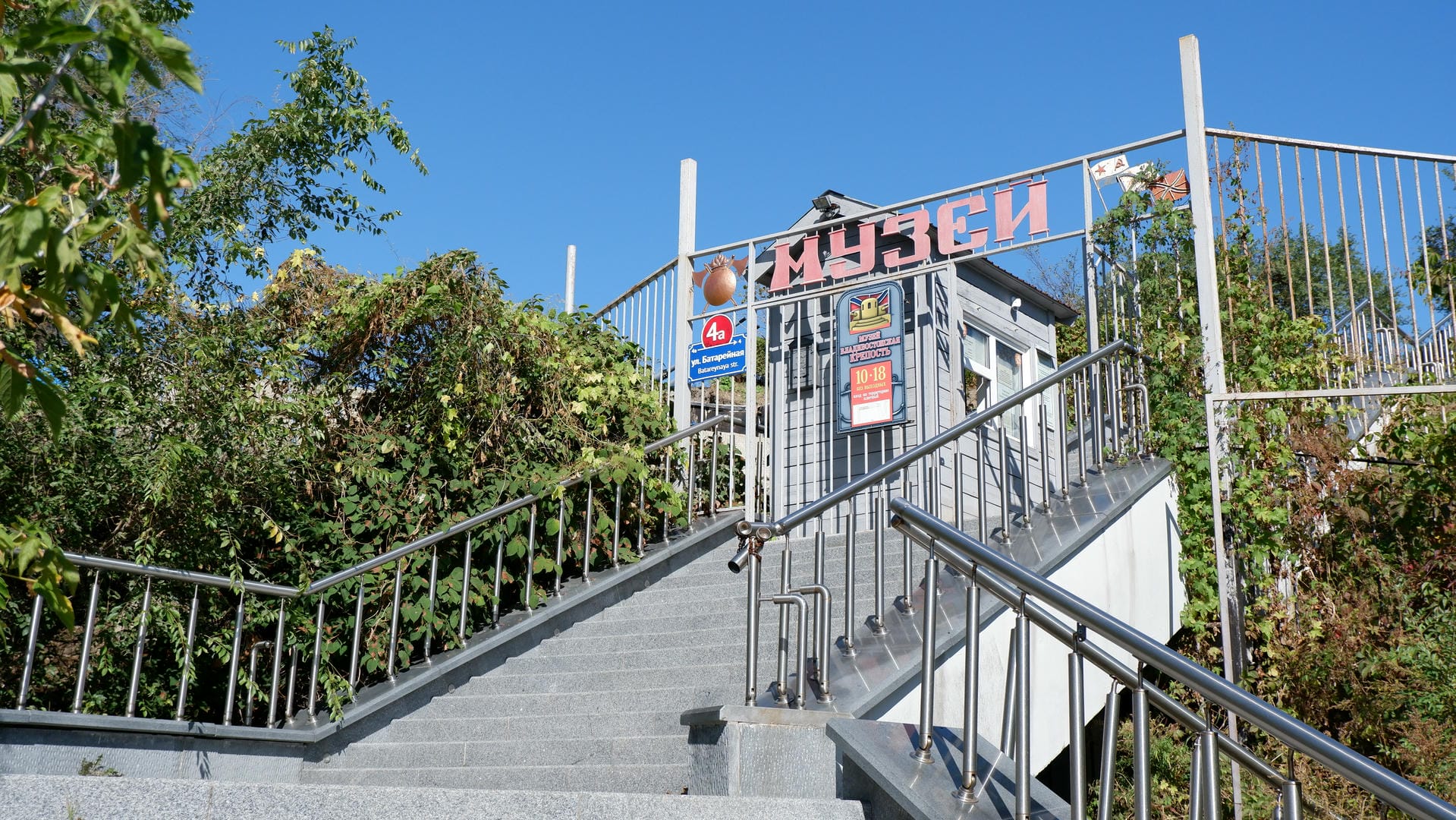 The entrance of a museum with stairs and metal railings, surrounded by green plants.