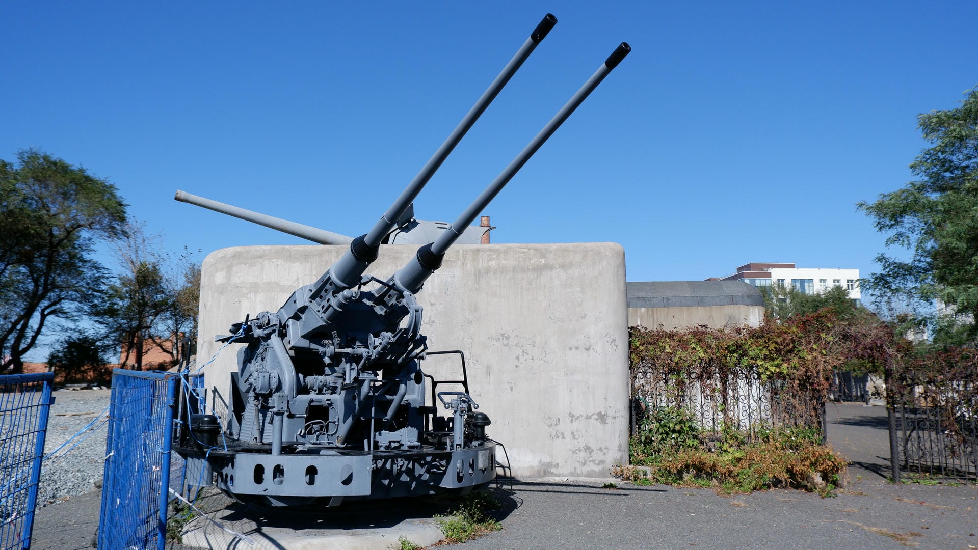 Two anti-aircraft guns installed side by side on a concrete base, with a blue fence nearby.