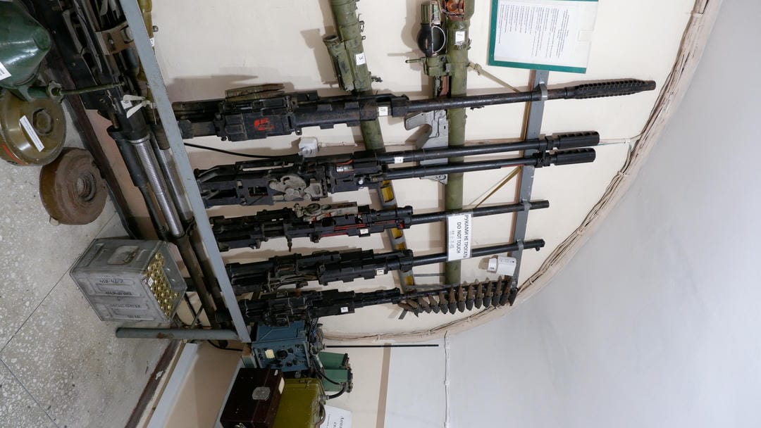 Several rocket launchers and other military equipment hang on the wall, alongside boxes and cans.