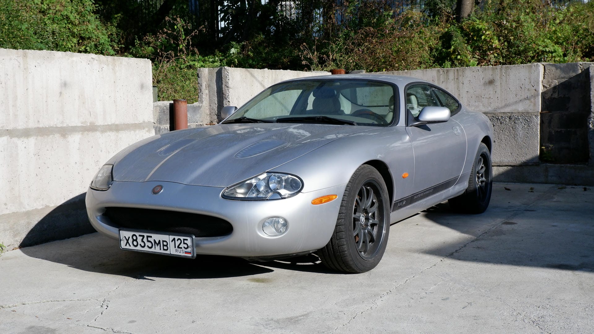 A silver Jaguar sports car parked in front of a concrete wall, with license plate number X835MB 125.