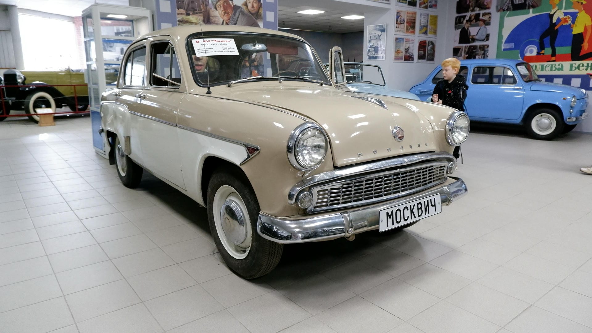A beige vintage car parked inside an exhibition hall, with license plate МОСКВИЧ.