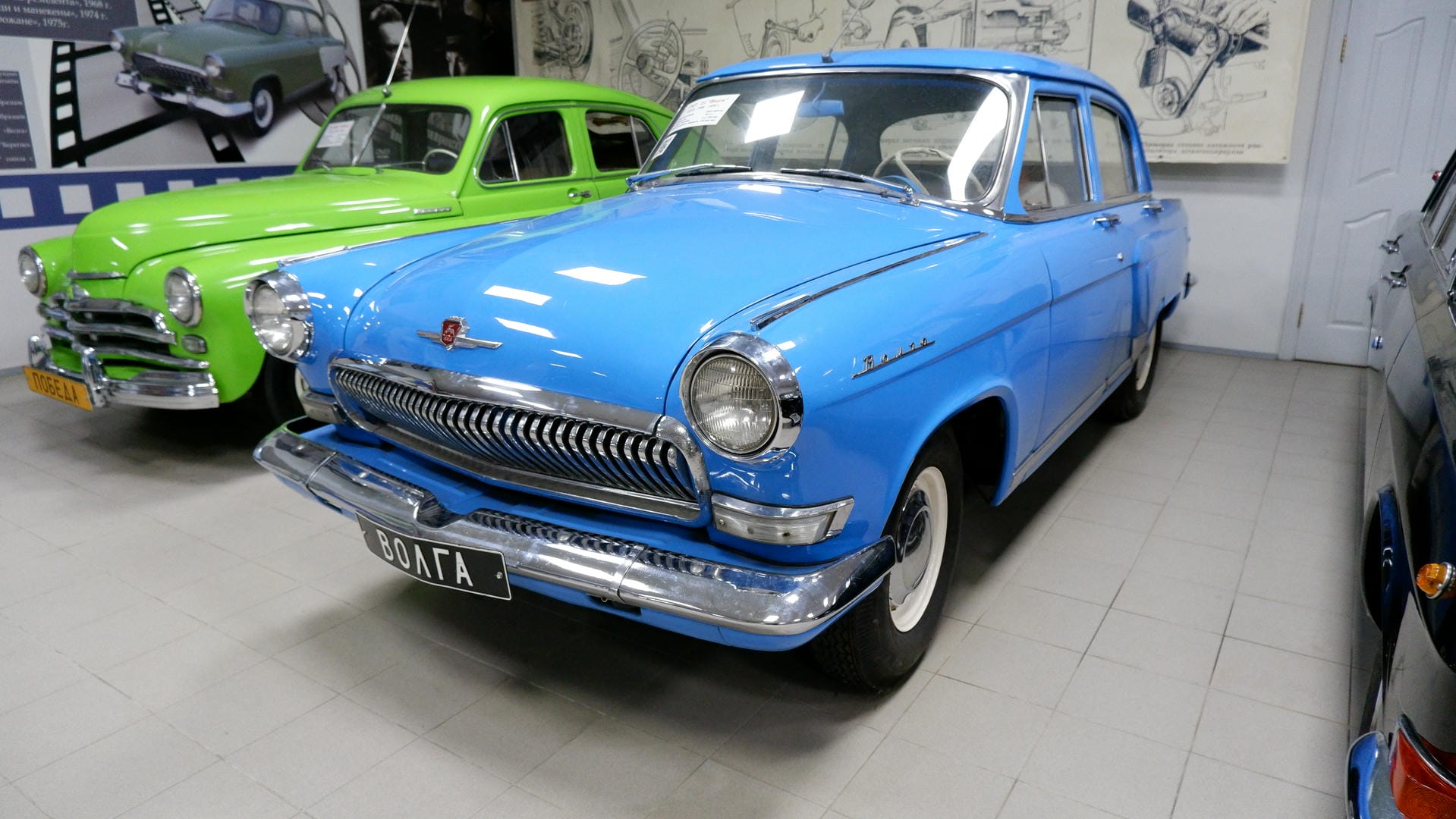 A blue vintage car parked inside an exhibition hall, with license plate ВОЛГА.