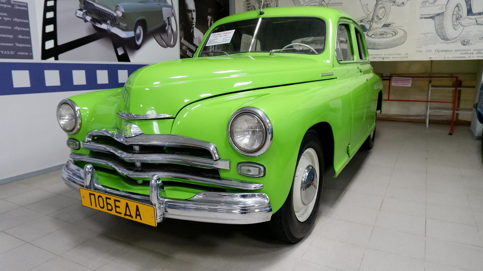 A green retro car with license plate “ПОБЕДА,” parked in an indoor exhibition space.