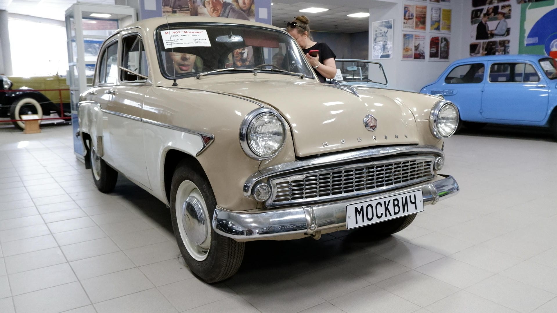 A beige retro car with license plate “МОСКВИЧ,” parked in an indoor exhibition space.