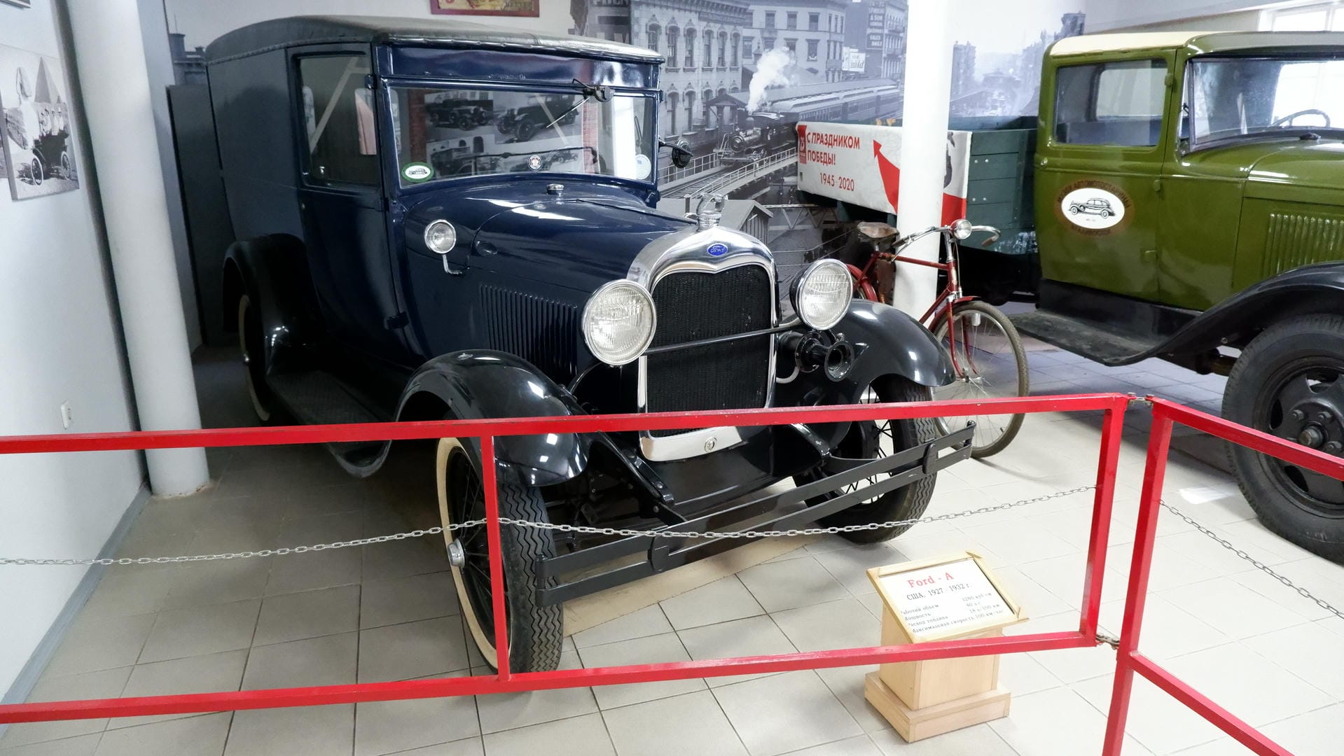 A black vintage car with a red railing in front, next to a wooden signboard.