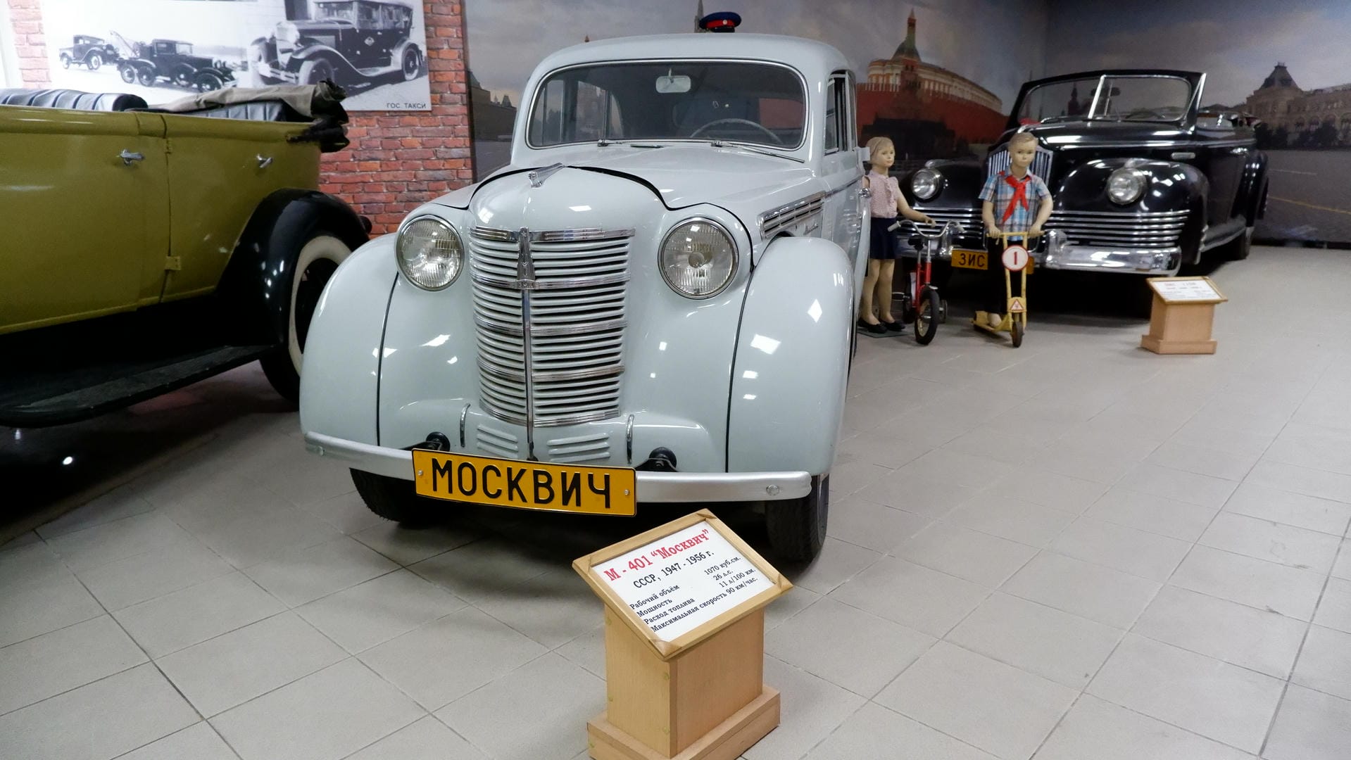 A light blue vintage car with license plate “МОСКВИЧ,” accompanied by two mannequins dressed in retro clothing.