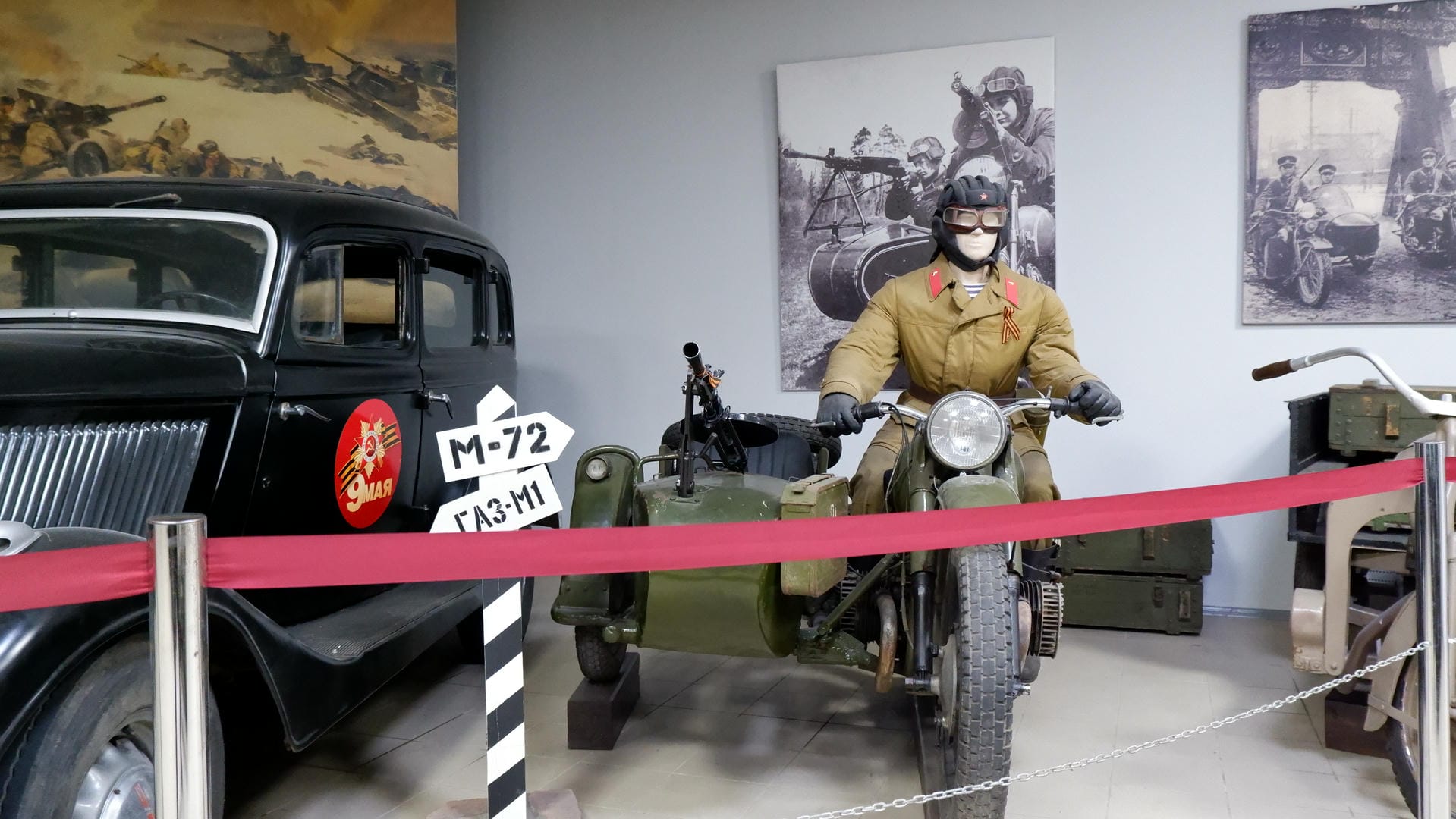 A military dummy in uniform riding a motorcycle, with a black car beside it, against a backdrop of a war scene painting.