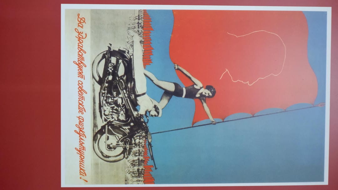 A poster showing one person riding a motorcycle while another stands on the back holding a flag, against a red flag background.