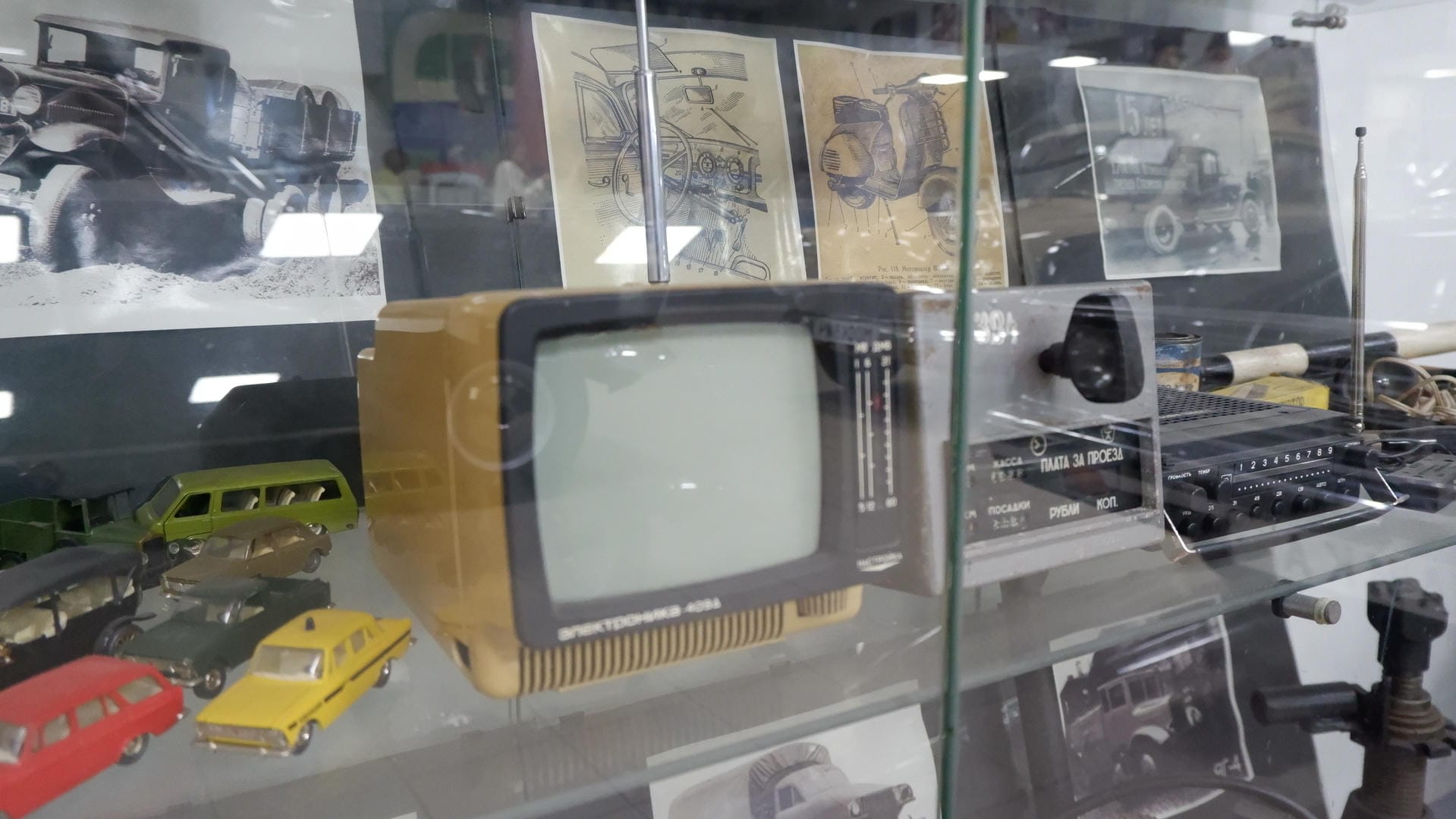 A display case containing an old television, radio, and other items, with car blueprints in the background.