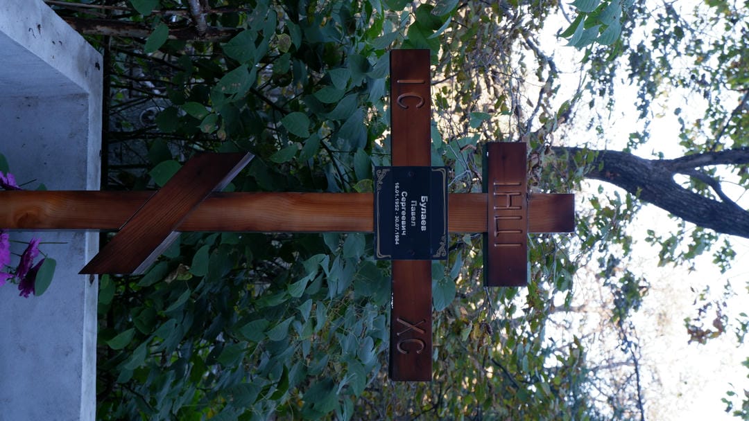 A wooden cross standing in a graveyard, with Russian inscriptions and a black nameplate on it.