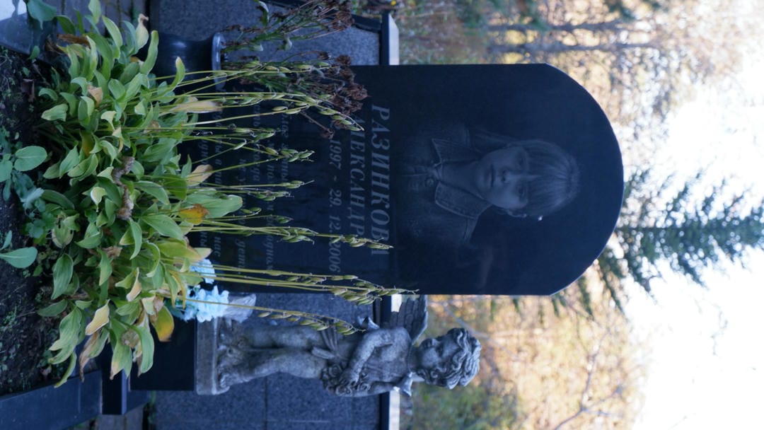 A black tombstone engraved with a young girl’s photo and name “Разинкова,” accompanied by a small angel statue.