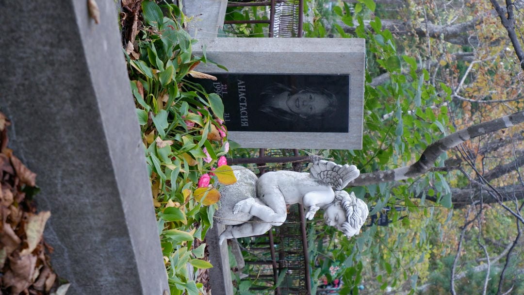 A gray tombstone engraved with a young girl’s photo and name “Анастасия,” accompanied by a small angel statue.