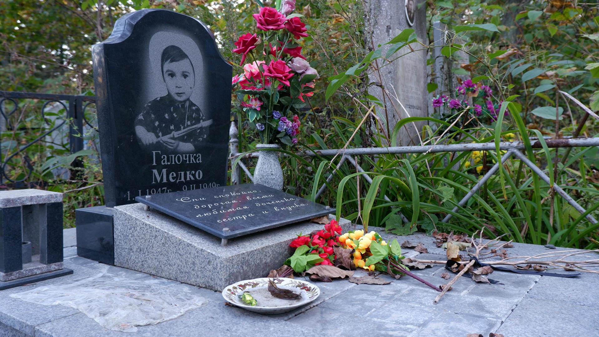 A black tombstone engraved with a young boy’s photo and name “Галочка Медко,” surrounded by flowers.