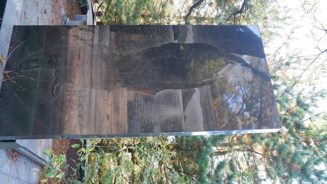 The tombstone is engraved with a male portrait, with trees and sky in the background.