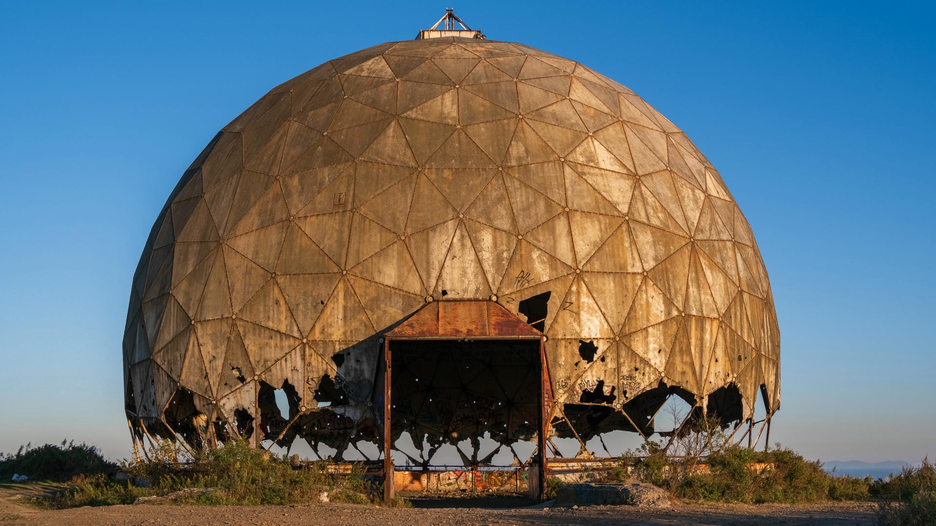 A dilapidated dome structure, its surface covered in rust and damage, with multiple openings at the base.