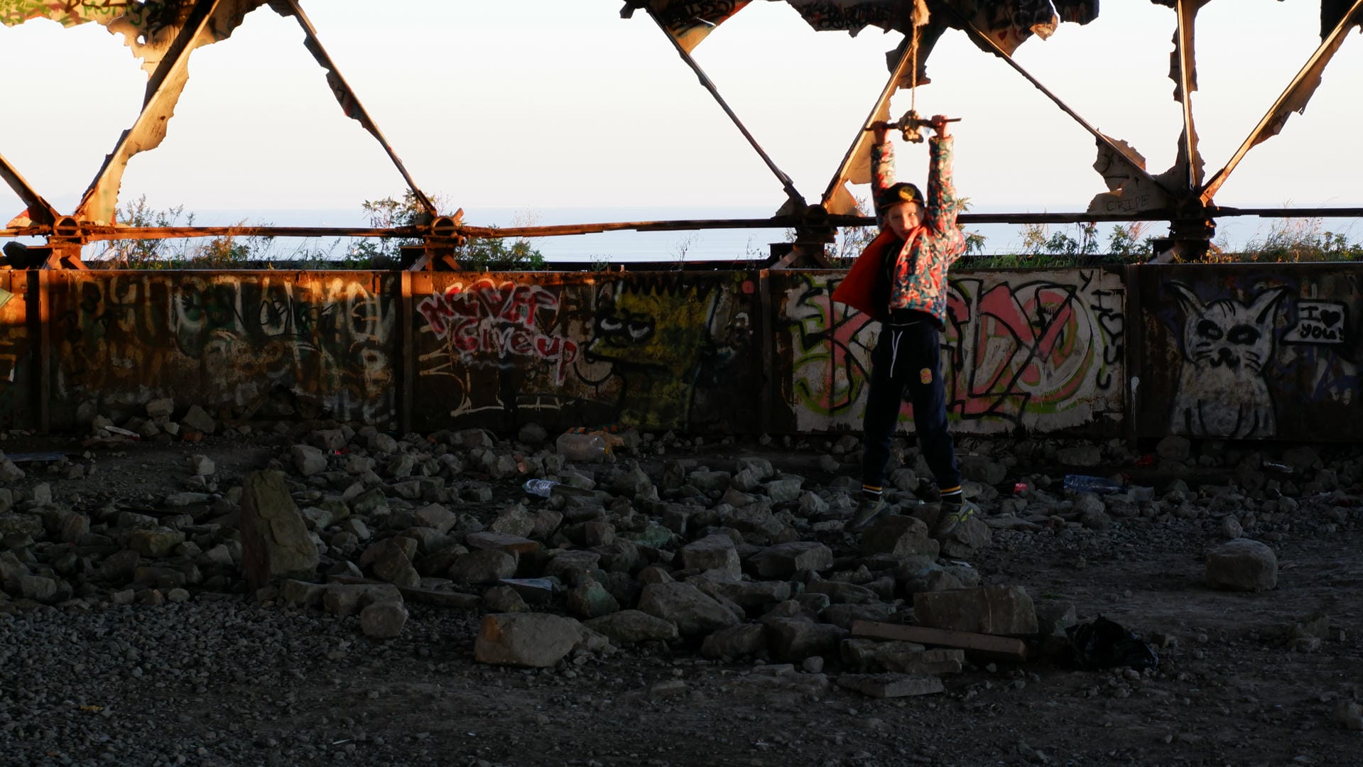 A person stands in the ruins with arms raised, with a graffiti wall and broken structures in the background.
