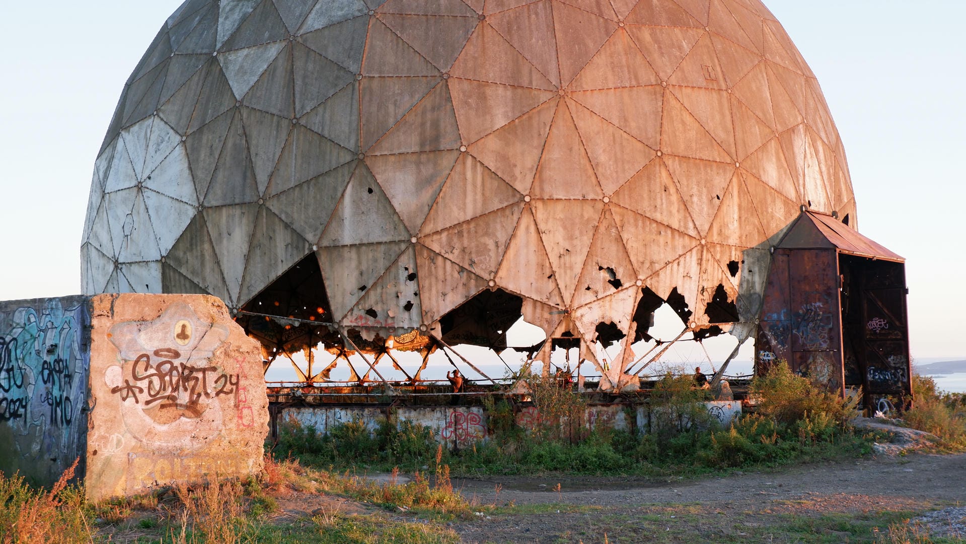 A large dome structure, visibly damaged and rusted, with a graffiti-covered stone tablet beside it.