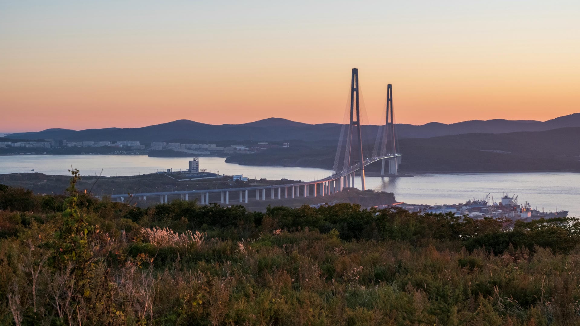 A bridge spans the bay as the sun sets, with an orange-yellow gradient sky. The bridge connects both shores, with mountains in the distance.