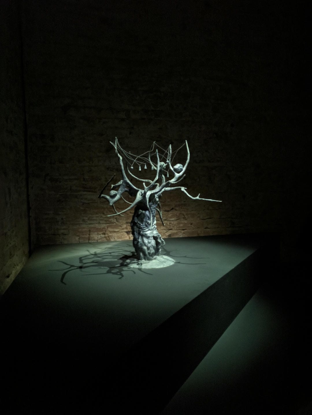 A twisted tree with ornaments hanging from its trunk and branches, with a brick wall in the background.