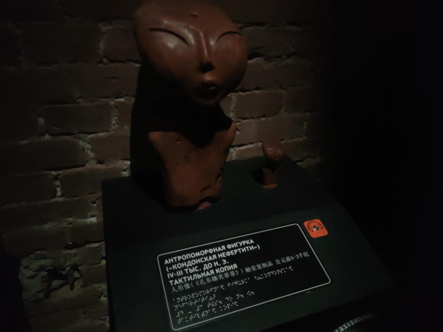 A brown human-shaped sculpture, armless, with a calm facial expression, against a brick wall.