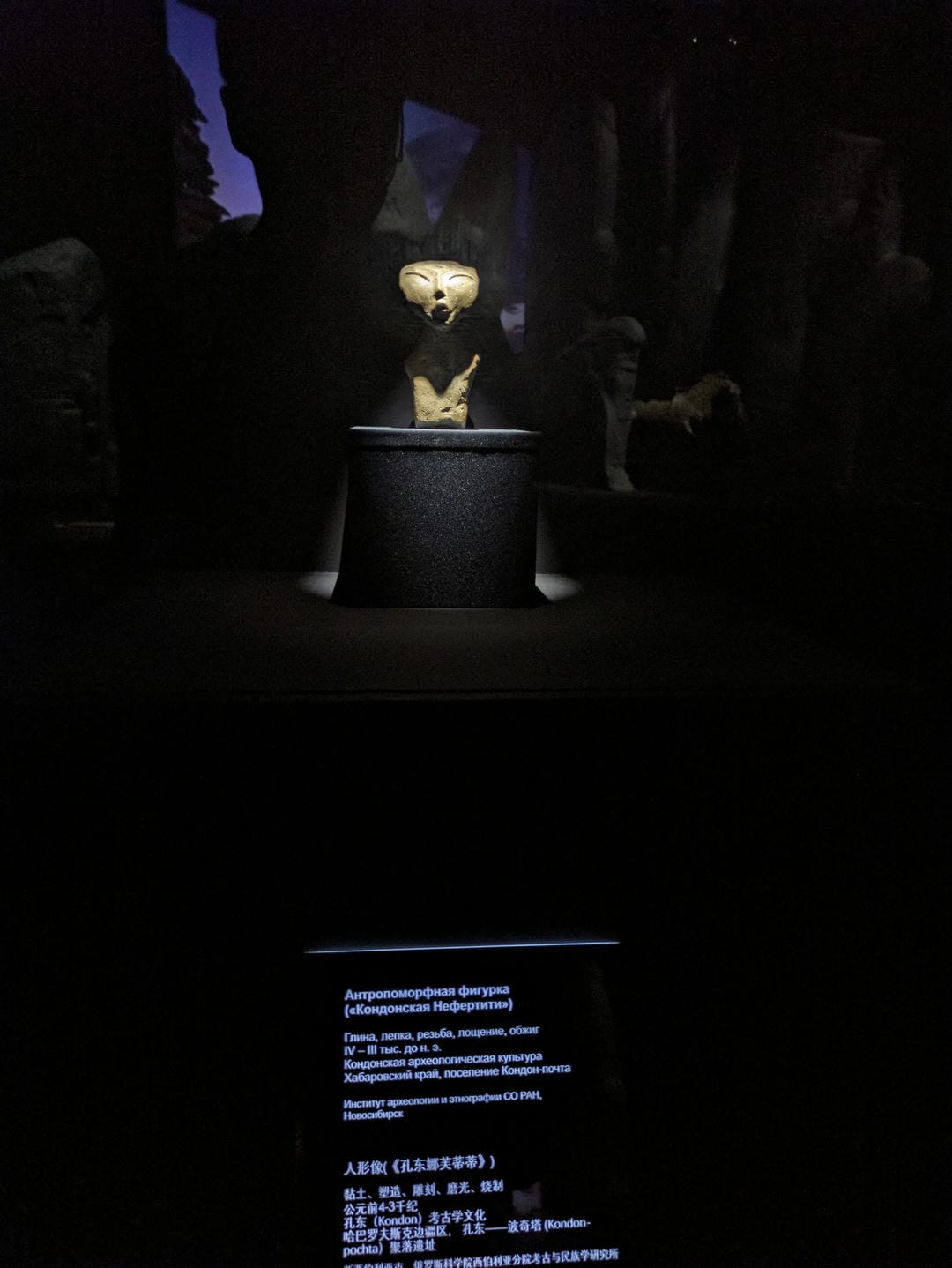 A human-shaped sculpture with a large head, closed eyes, slightly open mouth, and a dark background.