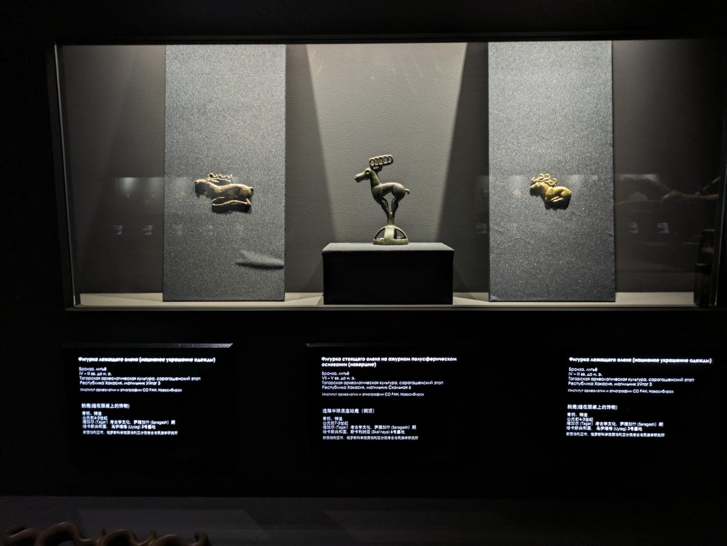 Three bronze objects: a lying animal on the left, a standing animal in the middle, and another lying animal on the right, against a gray background.