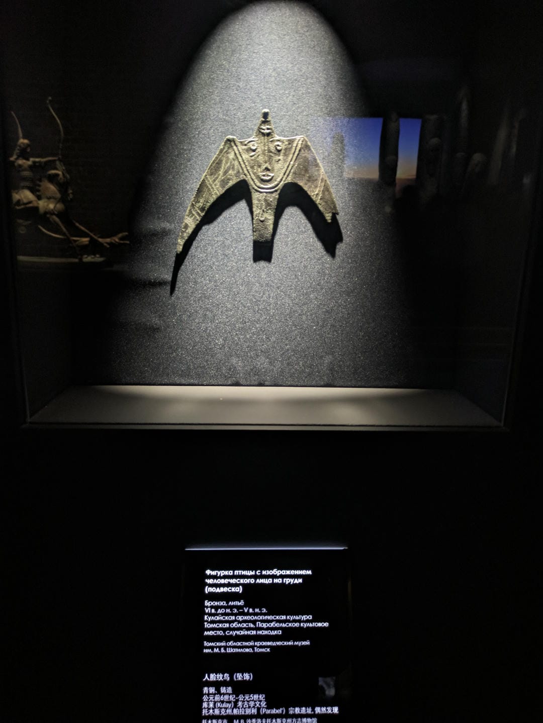 A bird-shaped pendant with a human face design, facing forward with wings spread, against a black background.