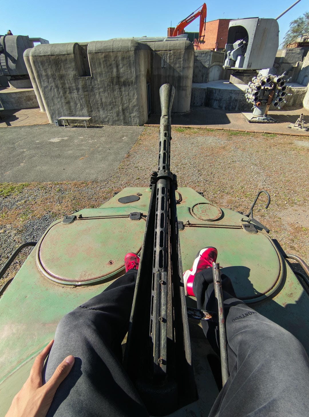 A person sitting on a tank, holding a machine gun, with concrete structures and military equipment in the background.