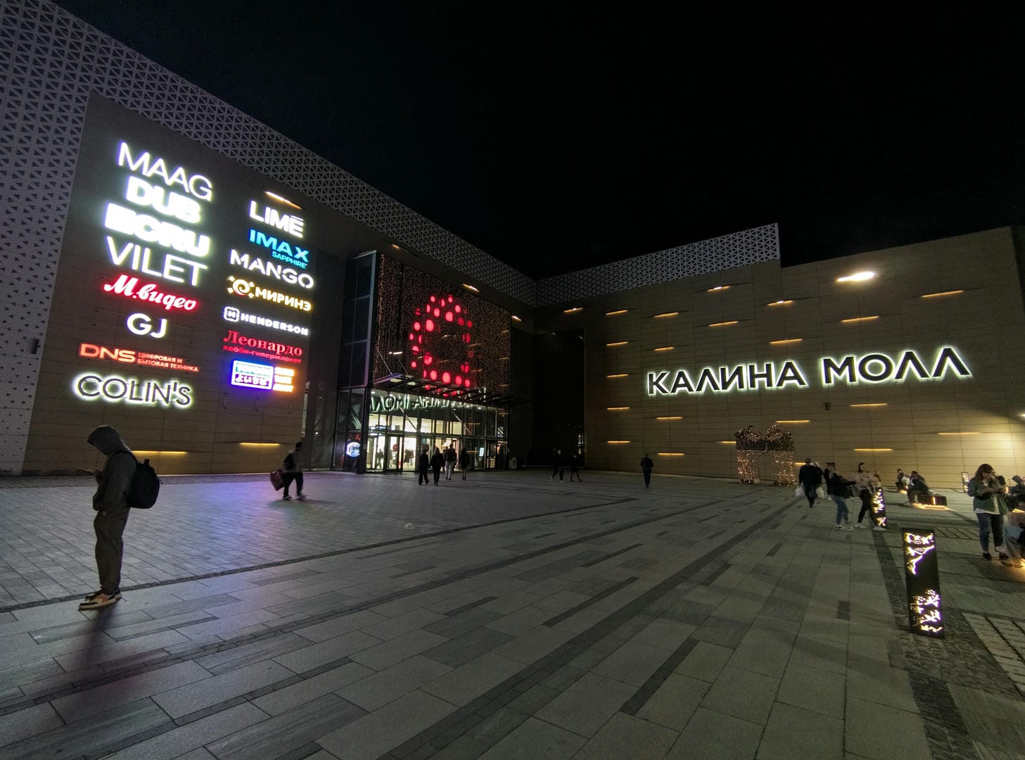 At night, many store signs are lit up in a city square where people are walking around.