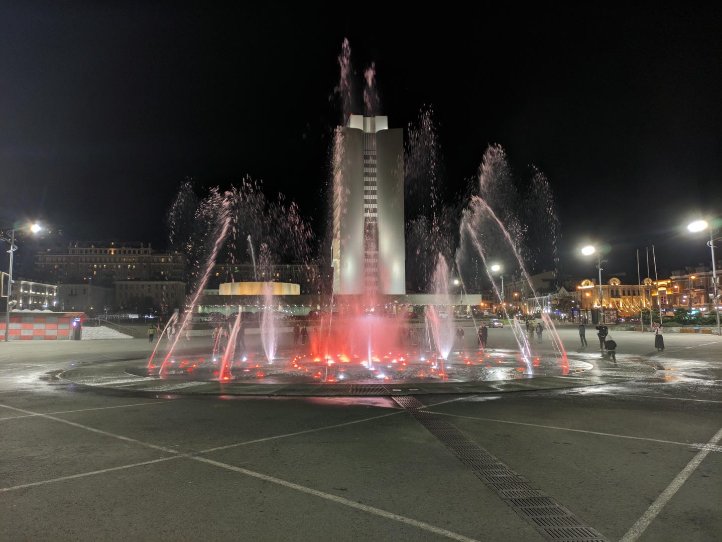 In the center of a nighttime square, there is a fountain with water jets dyed red by lights.