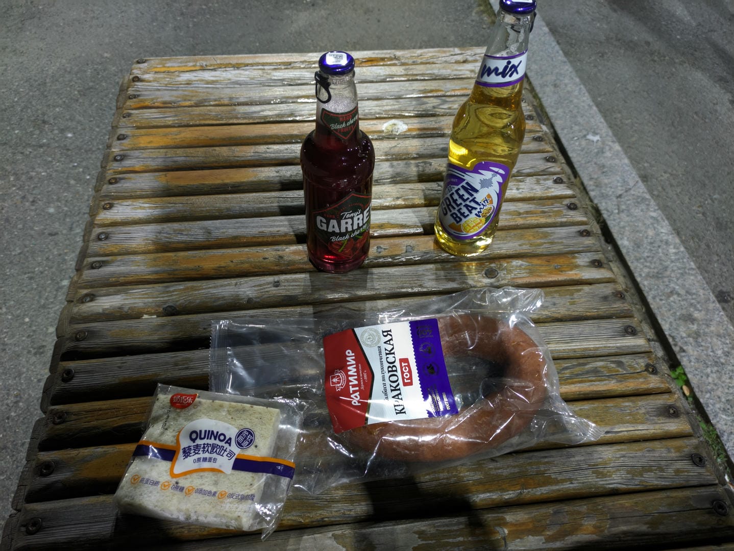 On a wooden table, there are two bottles of drinks and two types of food. One bottle is red Garrafa Black drink, and the other is yellow Green Bean Mix drink. The packaging of the food on the left says “QUINOA,” and the one on the right is a round sausage.