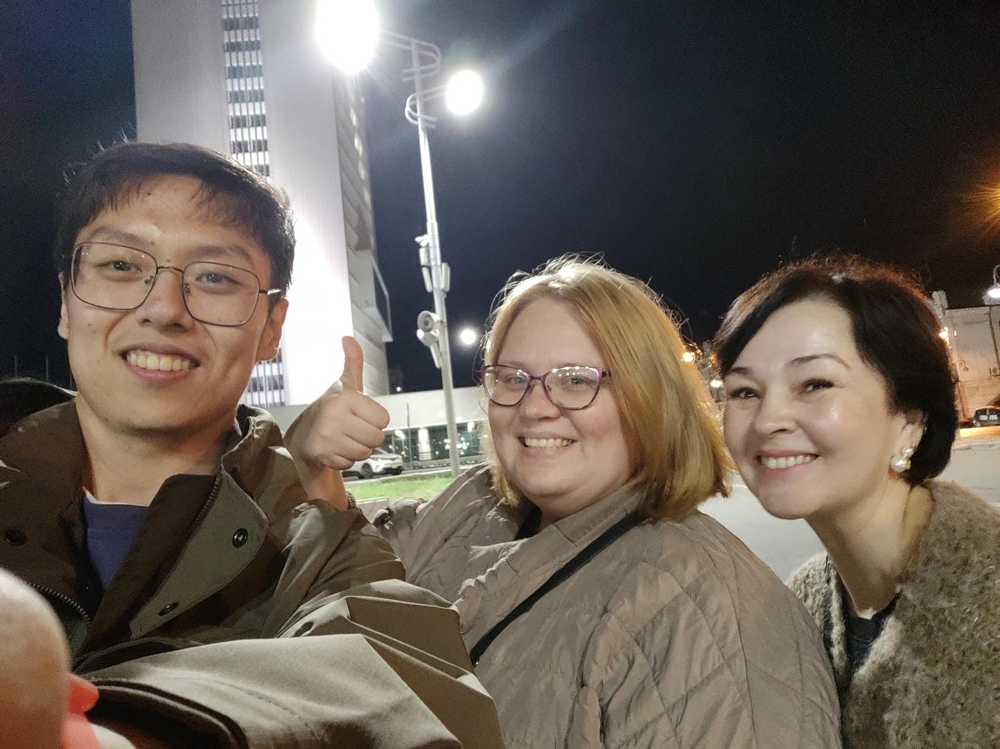 Three people taking a group photo outdoors at night. The person on the left is wearing glasses and a brown coat, giving a thumbs-up. The person in the middle is wearing transparent glasses and a light-colored coat, smiling. The person on the right has short hair, wears a dark coat, and also smiles. Buildings and streetlights can be seen in the background.