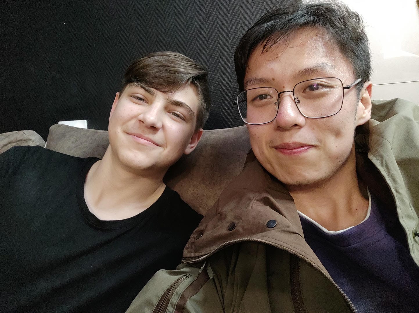 Two people sitting on a sofa, the person on the left wearing a black top, and the person on the right wearing glasses and a brown coat.